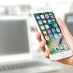 Close-up view of a smartphone displaying apps, held by a hand, with a blurred laptop in the background., tags: apples digital markets - pexel