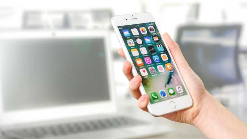 Close-up view of a smartphone displaying apps, held by a hand, with a blurred laptop in the background., tags: apples digital markets - pexel